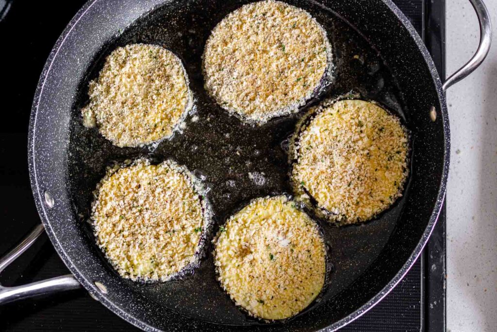 Breaded eggplant slices frying in oil in a skillet.