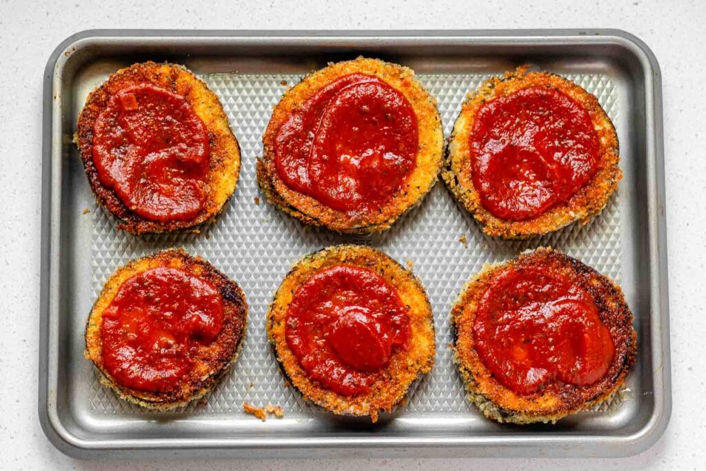 Breaded eggplant slices on a sheet pan with tomato sauce on top.