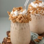 A milkshake in a glass jar topped with whipped cream, cereal and a glass straw.