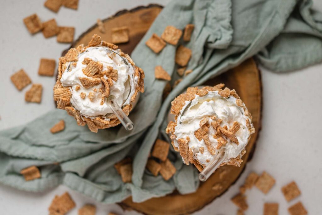 Top down view of two milkshakes topped with whipped cream and cereal on a wooden board with a light green cloth.