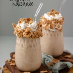 A milkshake in a glass jar topped with whipped cream and cereal on a wooden board with black text above.