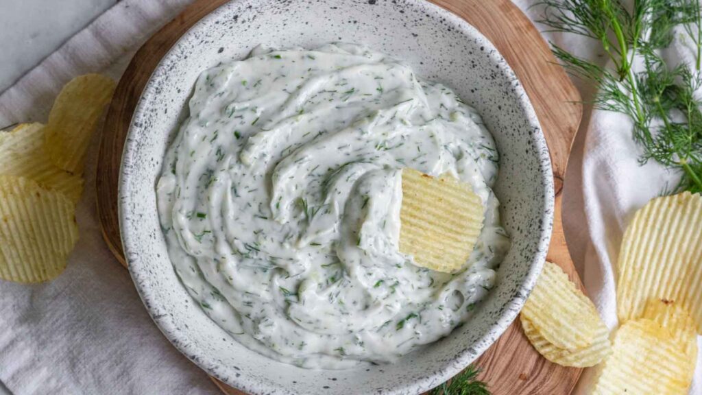 White dip in bowl with herbs in it, with potato chips to dip.