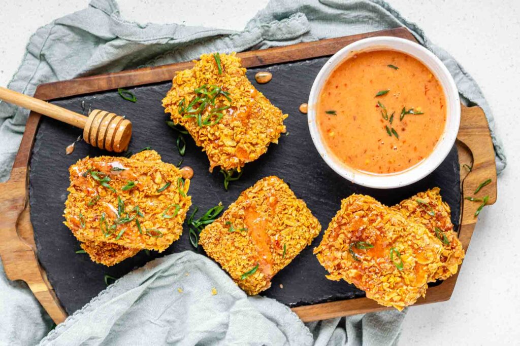 Breaded tofu on a board with a bowl of hot honey.