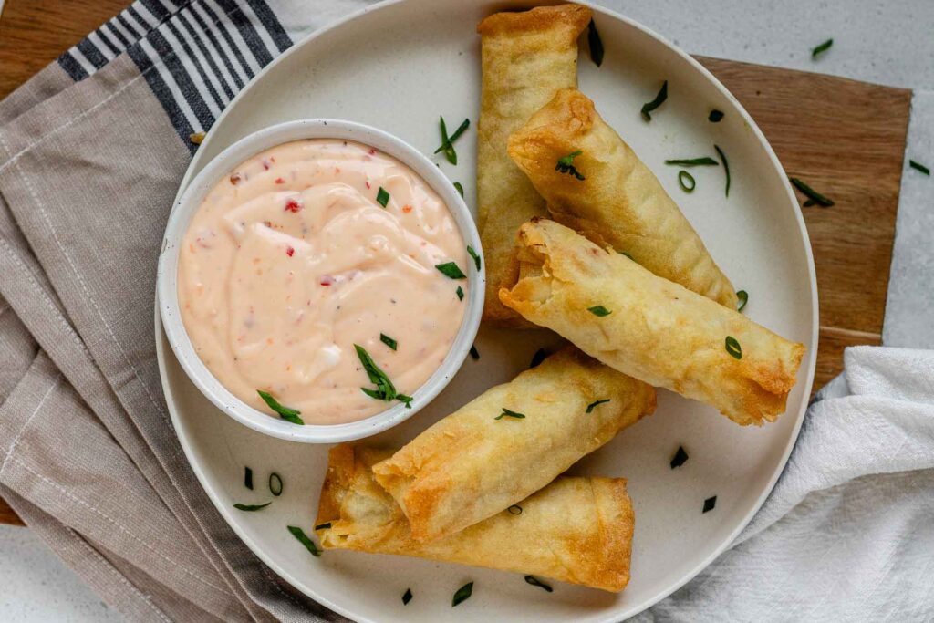 Sweet chili mayo in a bowl on a plate beside spring rolls.