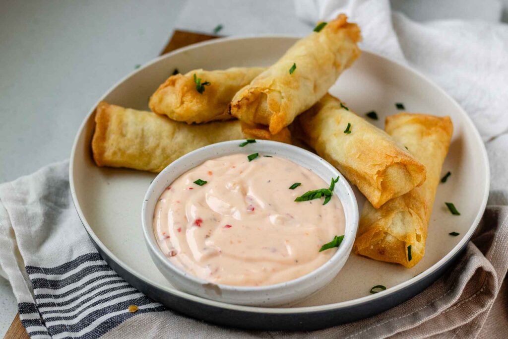 Sweet chili mayo in a bowl on a plate beside spring rolls.