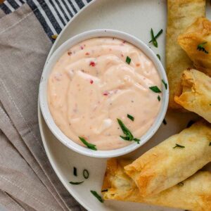 Sweet chili mayo in a bowl on a plate beside spring rolls.